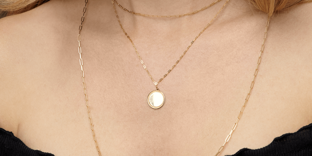Close-up of a woman wearing layered gold chains with a small round pendant over a black off-shoulder top.