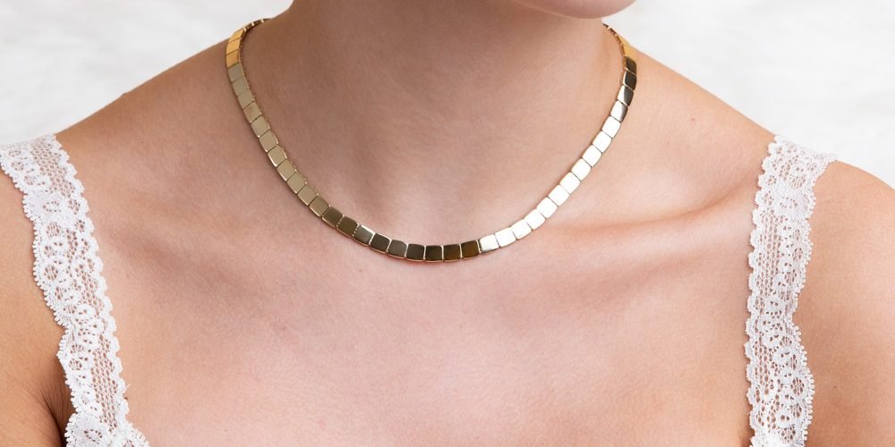Close-up of a woman wearing a gold square-link choker necklace with a white lace-trimmed top.